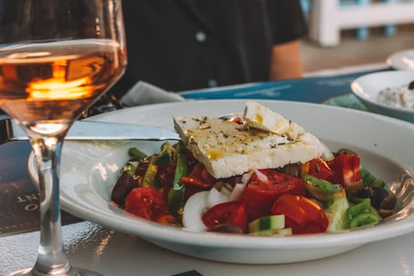 A plate of Greek salad with chopped cucumbers, tomatoes, onions, olives, and a large slab of feta cheese drizzled with olive oil is served on a table. Beside the plate is a glass of rose wine, partially filled, catching the light.