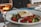 A plate of Greek salad with chopped cucumbers, tomatoes, onions, olives, and a large slab of feta cheese drizzled with olive oil is served on a table. Beside the plate is a glass of rose wine, partially filled, catching the light.