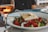 A plate of Greek salad with chopped cucumbers, tomatoes, onions, olives, and a large slab of feta cheese drizzled with olive oil is served on a table. Beside the plate is a glass of rose wine, partially filled, catching the light.