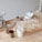 Various kitchen items are placed on a wooden countertop. There is a pasta machine, a metal cup, a white mixing bowl with a spoon, a glass container filled with flour, and a small jar with nuts. Circular dough pieces are scattered on a floured surface, suggesting preparations for a meal.