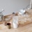 Various kitchen items are placed on a wooden countertop. There is a pasta machine, a metal cup, a white mixing bowl with a spoon, a glass container filled with flour, and a small jar with nuts. Circular dough pieces are scattered on a floured surface, suggesting preparations for a meal.
