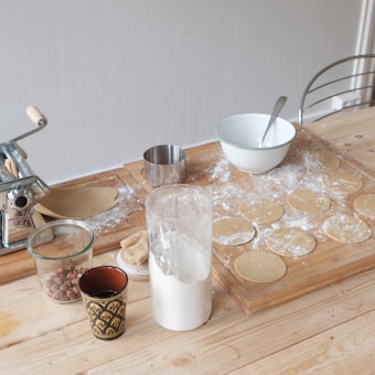 Various kitchen items are placed on a wooden countertop. There is a pasta machine, a metal cup, a white mixing bowl with a spoon, a glass container filled with flour, and a small jar with nuts. Circular dough pieces are scattered on a floured surface, suggesting preparations for a meal.