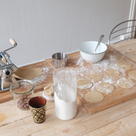 Various kitchen items are placed on a wooden countertop. There is a pasta machine, a metal cup, a white mixing bowl with a spoon, a glass container filled with flour, and a small jar with nuts. Circular dough pieces are scattered on a floured surface, suggesting preparations for a meal.