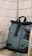 A sleek, modern backpack with a roll-top design is positioned against a woven rattan background. It features black straps and buckles with a light gray body, creating a contrast in textures. A small green plant adds a touch of nature to the setting.