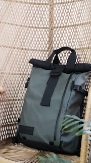 A sleek, modern backpack with a roll-top design is positioned against a woven rattan background. It features black straps and buckles with a light gray body, creating a contrast in textures. A small green plant adds a touch of nature to the setting.