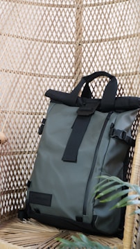 A sleek, modern backpack with a roll-top design is positioned against a woven rattan background. It features black straps and buckles with a light gray body, creating a contrast in textures. A small green plant adds a touch of nature to the setting.