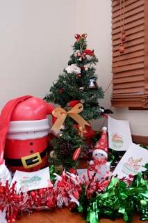 A small, decorated Christmas tree is adorned with festive ornaments, surrounded by holiday-themed crafts and decorations, including tinsel in red and green hues. A Santa-shaped container and a small snowman figurine wearing a red hat are also present. The scene is set against a backdrop of wooden blinds.