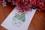 A hand-drawn illustration features a cupcake decorated to resemble a Christmas tree, adorned with colorful ornaments and lights. A small Santa figure is perched on the top, smiling beneath a tinsel and star ornament. The image is placed on a wooden surface, accompanied by red and white tinsel and paper stars.