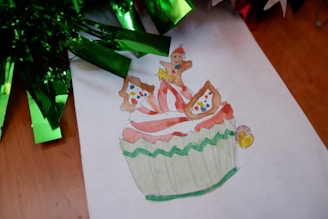 A watercolor illustration of a festive cupcake with red and white swirled frosting topped with small gingerbread figures. The cupcake wrapper features a green zigzag pattern. Next to the illustration, shiny green foil decorations are partially visible on a wooden surface.