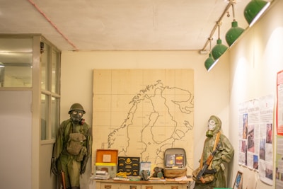 A room displaying various military and historical artifacts. Two mannequins are dressed in military gear and gas masks, standing on either side of a table. Behind them is a large map of a geographical area pinned to the wall. The table is filled with memorabilia, including books, a television, and other items. The room has a white ceiling with green light fixtures hanging down on the right side.