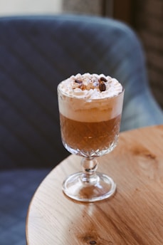 A glass goblet filled with a brown beverage topped with whipped cream and coffee beans. It rests on a round wooden table with a quilted blue chair in the background.