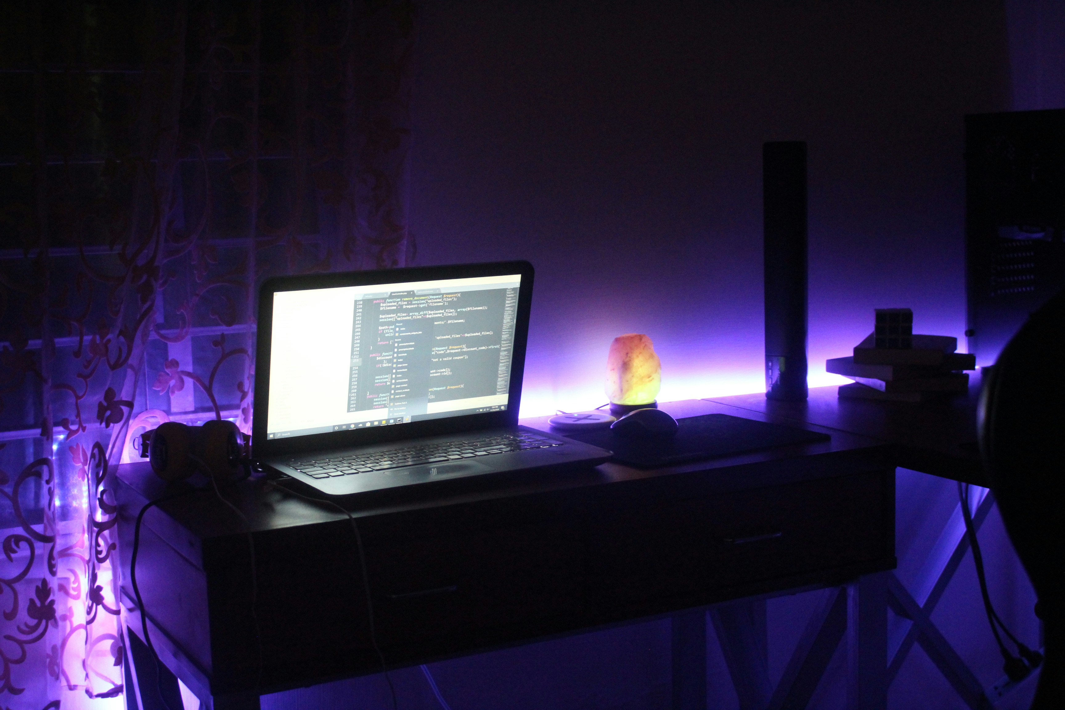 Laptop with code on dark desk, warm atmospheric lamp, moody dim room, dark wooden surface, deep shadows