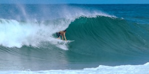 A surfer skillfully rides inside a large, powerful wave, with water crashing around. The wave is curling over the surfer, creating a dramatic and energetic scene as mist sprays into the air.