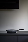 A black non-stick frying pan is placed on a dark countertop beneath a large, dark exhaust hood. The background is a plain, light-colored wall.