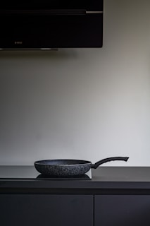 A black non-stick frying pan is placed on a dark countertop beneath a large, dark exhaust hood. The background is a plain, light-colored wall.