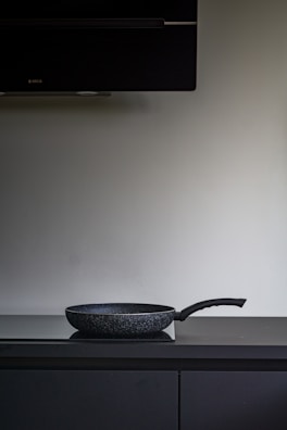 A black non-stick frying pan is placed on a dark countertop beneath a large, dark exhaust hood. The background is a plain, light-colored wall.
