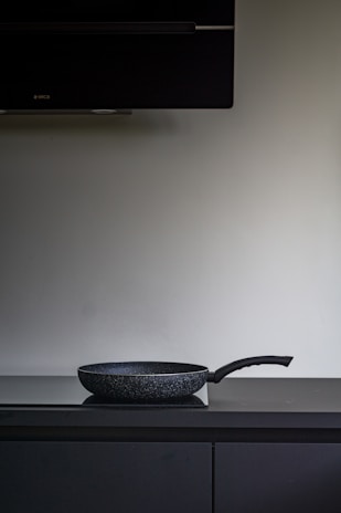 A black non-stick frying pan is placed on a dark countertop beneath a large, dark exhaust hood. The background is a plain, light-colored wall.