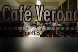 The image depicts the interior of a café viewed through a glass window with the words 'Café Verona' prominently displayed. Inside, a person is seen holding a tray, surrounded by plates and restaurant equipment. The setting is dimly lit, with warm lighting creating a cozy atmosphere.