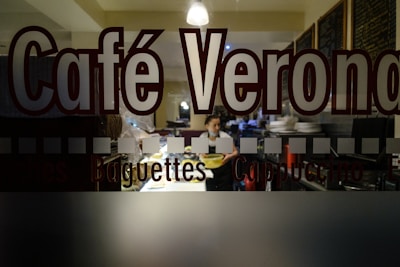 The image depicts the interior of a café viewed through a glass window with the words 'Café Verona' prominently displayed. Inside, a person is seen holding a tray, surrounded by plates and restaurant equipment. The setting is dimly lit, with warm lighting creating a cozy atmosphere.