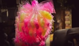 A bunch of cotton candy packaged in transparent plastic bags, displaying vibrant colors in pink and yellow hues. The background is slightly blurred, highlighting the bright candy against darker surroundings.