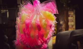A bunch of cotton candy packaged in transparent plastic bags, displaying vibrant colors in pink and yellow hues. The background is slightly blurred, highlighting the bright candy against darker surroundings.