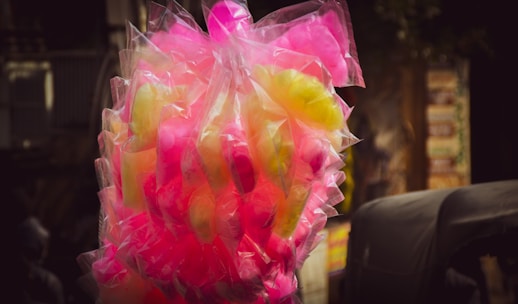 A bunch of cotton candy packaged in transparent plastic bags, displaying vibrant colors in pink and yellow hues. The background is slightly blurred, highlighting the bright candy against darker surroundings.