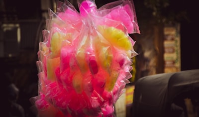 A bunch of cotton candy packaged in transparent plastic bags, displaying vibrant colors in pink and yellow hues. The background is slightly blurred, highlighting the bright candy against darker surroundings.