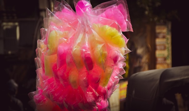 A bunch of cotton candy packaged in transparent plastic bags, displaying vibrant colors in pink and yellow hues. The background is slightly blurred, highlighting the bright candy against darker surroundings.