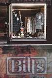 A vintage-style shop window display showcases an array of men's clothing, including two formal coats placed on mannequins. Various patterned shirts hang neatly on a rack in the background. Accessories such as ties and handkerchiefs add a vibrant touch to the scene. A name sign, 'Bill's', is visible at the bottom of the image near a textured street barrier.
