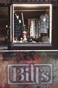 A vintage-style shop window display showcases an array of men's clothing, including two formal coats placed on mannequins. Various patterned shirts hang neatly on a rack in the background. Accessories such as ties and handkerchiefs add a vibrant touch to the scene. A name sign, 'Bill's', is visible at the bottom of the image near a textured street barrier.