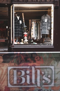 A vintage-style shop window display showcases an array of men's clothing, including two formal coats placed on mannequins. Various patterned shirts hang neatly on a rack in the background. Accessories such as ties and handkerchiefs add a vibrant touch to the scene. A name sign, 'Bill's', is visible at the bottom of the image near a textured street barrier.