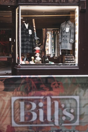 A vintage-style shop window display showcases an array of men's clothing, including two formal coats placed on mannequins. Various patterned shirts hang neatly on a rack in the background. Accessories such as ties and handkerchiefs add a vibrant touch to the scene. A name sign, 'Bill's', is visible at the bottom of the image near a textured street barrier.