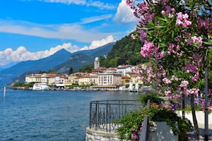A picturesque lakeside town with colorful buildings nestled against a backdrop of lush mountains. Pink flowers frame the right side, adding vibrant contrast to the blue sky and calm waters of the lake. A small pier with boats is visible along the shoreline, enhancing the idyllic scenery.