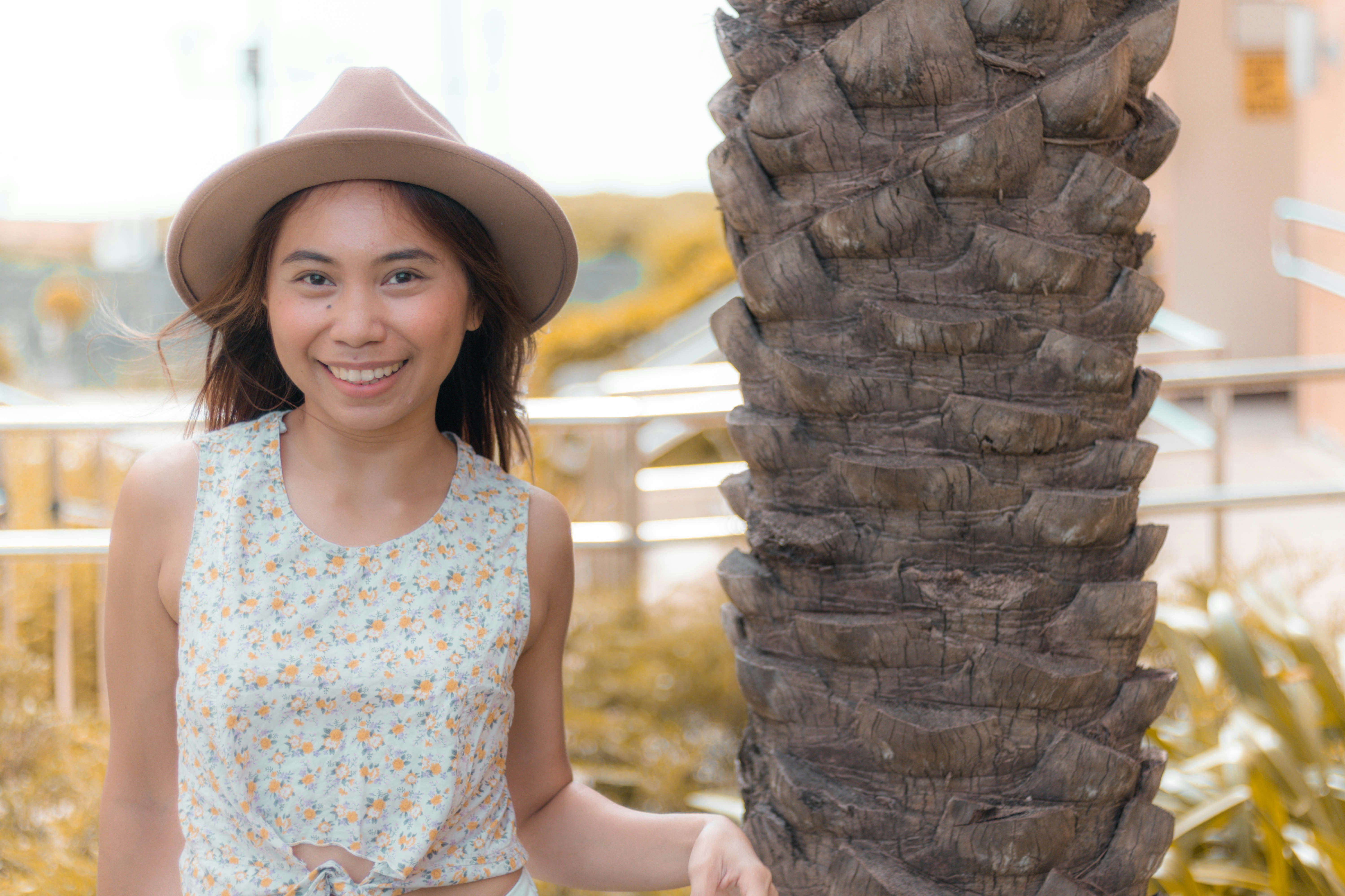Ragazza in canotta bianca e blu che indossa un cappello marrone in piedi accanto all'albero marrone durante il giorno