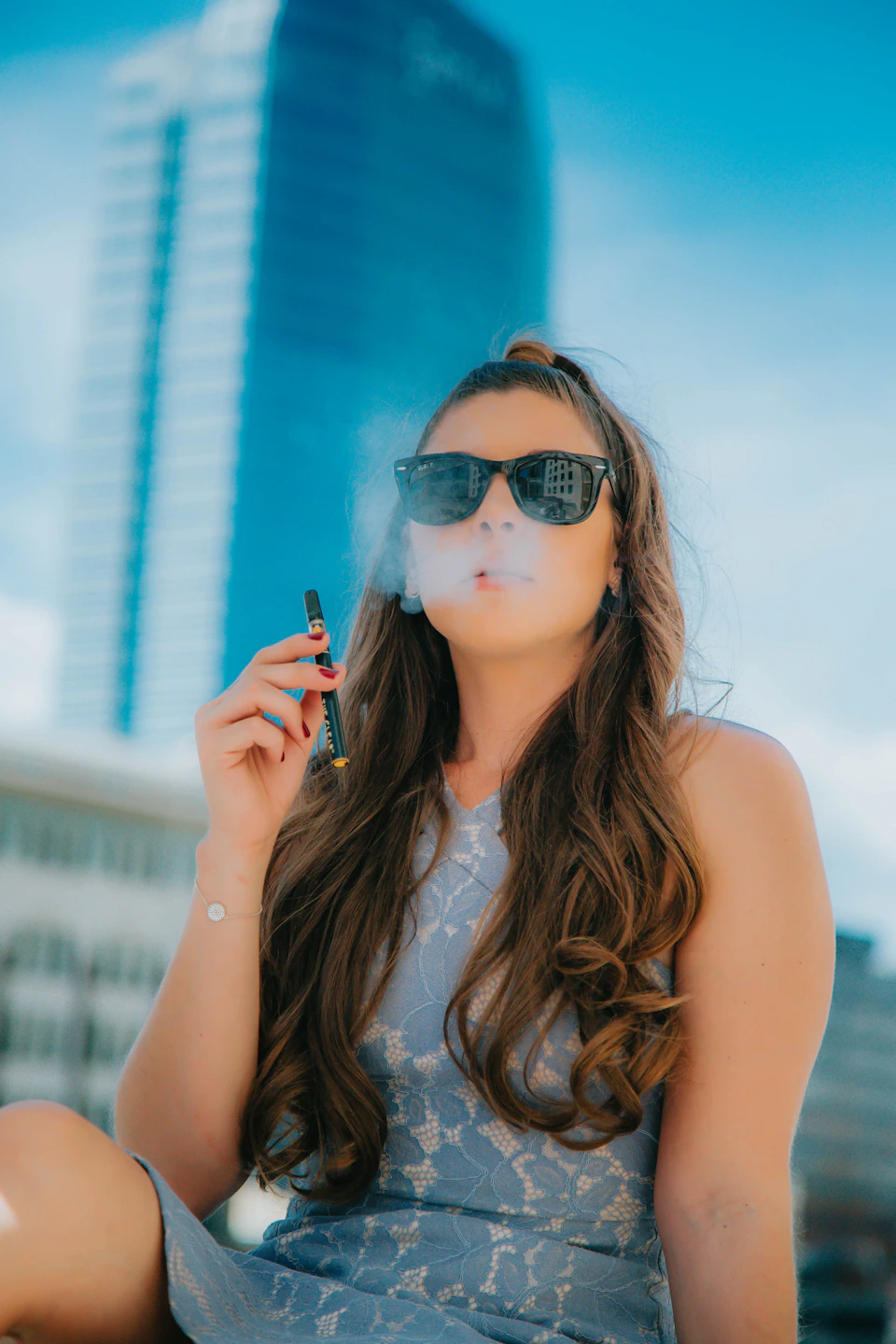Young woman sits on a bench, smoking an electronic cigarette with sunglasses on