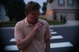 A person wearing a light-colored shirt and sunglasses is standing on a street with a zebra crossing, holding and using an electronic cigarette with a small light visible at the tip. The background shows suburban houses and some greenery, with the scene occurring during dusk or early evening.