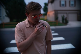 A person wearing a light-colored shirt and sunglasses is standing on a street with a zebra crossing, holding and using an electronic cigarette with a small light visible at the tip. The background shows suburban houses and some greenery, with the scene occurring during dusk or early evening.