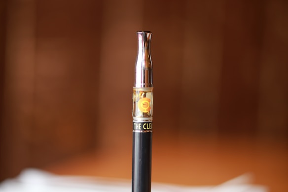 A vape pen with a metallic top and a transparent cartridge s filled with a liquid in the center of the frame. The background is softly blurred with brown tones, providing a warm and neutral context.