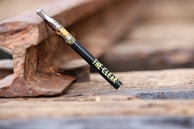 A vape pen labeled 'THE CLEAR' is resting diagonally against a rusty railroad track. The background features blurred wooden railroad ties and metal tracks, with a shallow depth of field focusing on the vape pen.