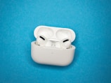 A pair of white wireless earbuds rests inside an open white charging case on a bright blue background. The earbuds have a sleek, modern design with black accents.