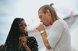 A man and a woman are standing close together, each holding a vape pen. The man is wearing a white shirt and has a watch on his wrist, while the woman is wearing a dark floral dress and yellow-tinted glasses. They appear engaged in their activity, with a blurred background suggesting an outdoor setting.