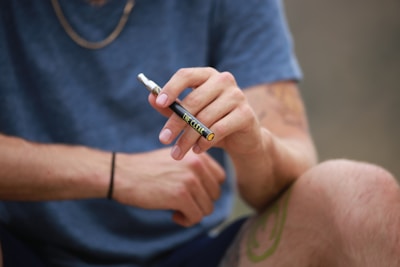 A close-up of a person holding a vape pen in their right hand. The individual is wearing a short-sleeved blue shirt and shorts, with a tattoo visible on their left arm and leg. A black wristband adorns the right wrist.