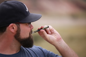 A man is holding a vape pen near his face. He is wearing a dark cap, glasses, and a t-shirt. The background is a blurred outdoor setting, suggesting a natural environment.
