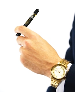 A hand holding a small black vape pen, with a golden watch on the wrist. The background is plain white, highlighting the hand and objects. The watch has a metallic strap and a round face.