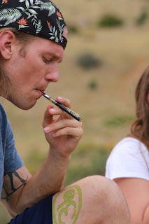 A person wearing a floral cap and a blue shirt is holding a vape pen to their mouth. Visible tattoos are on their arm and knee, and they are seated outdoors on a blurred natural background.