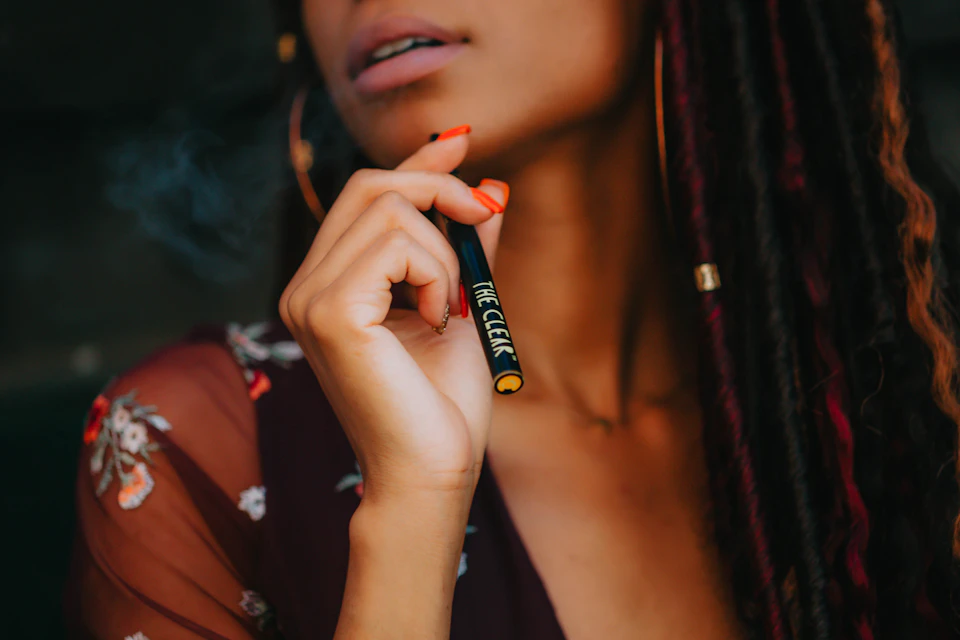 Woman's face in close-up with left hand holding black cigarette and right hand on necklace