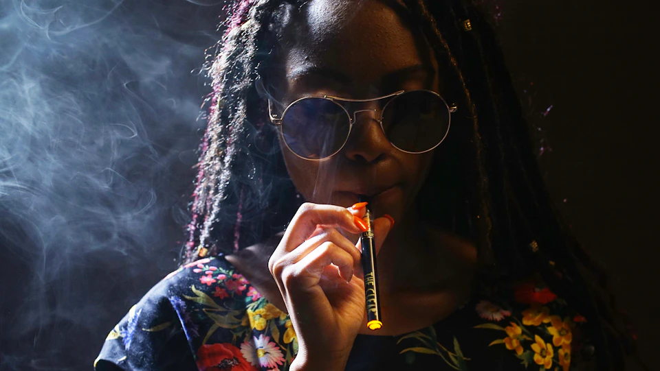 Person with dreadlocks wearing sunglasses holds an orange cigarette in their mouth against darkness