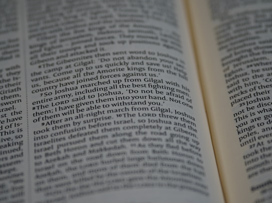 Close-up view of an open book with text in English, featuring words in different fonts, such as some words being italicized or bold. The focus is on lines involving a biblical text, suggesting a passage from scripture.