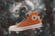 An orange Converse All Star high-top sneaker is prominently displayed against a blurred, nature-inspired backdrop. The shoe is detailed with black laces, a white rubber sole, and the iconic Converse logo on the side.