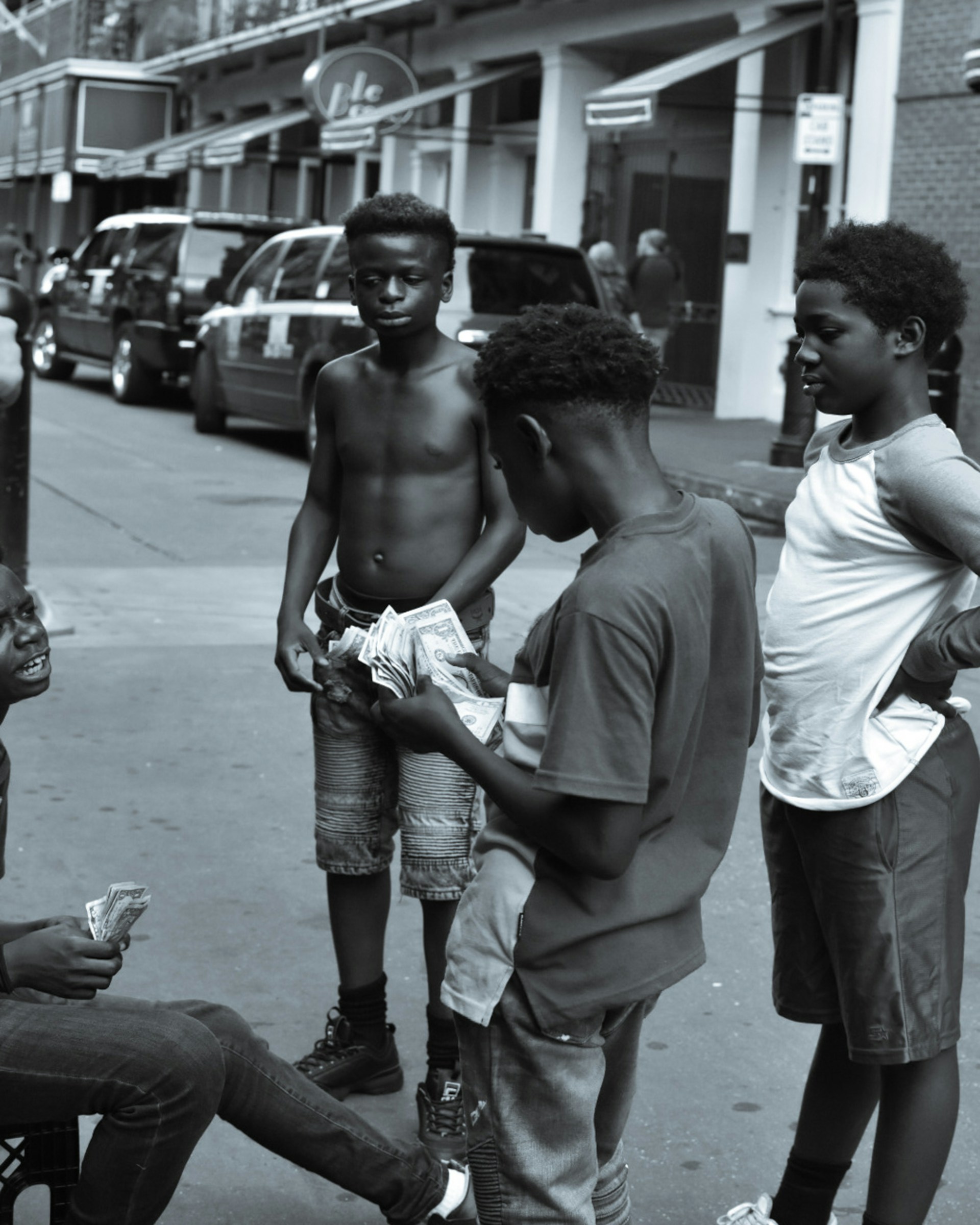 A group of four boys stands on an urban street. One boy is sitting and holding cash, while another boy holds a larger wad of cash. They seem to be engaged in a conversation with expressions that suggest a mixture of seriousness and curiosity. The background features parked cars and buildings.